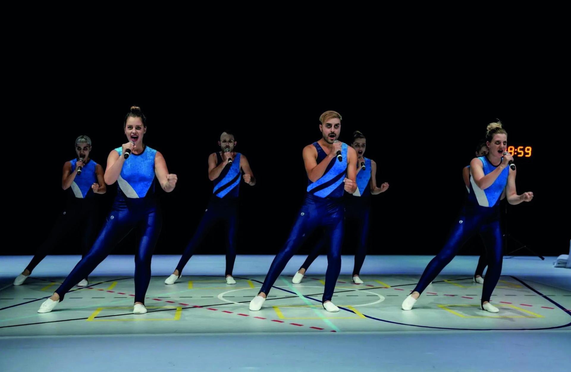 Six people in 80s tight-fitting gymnast outfits sing into a microphone and do gymnastics.