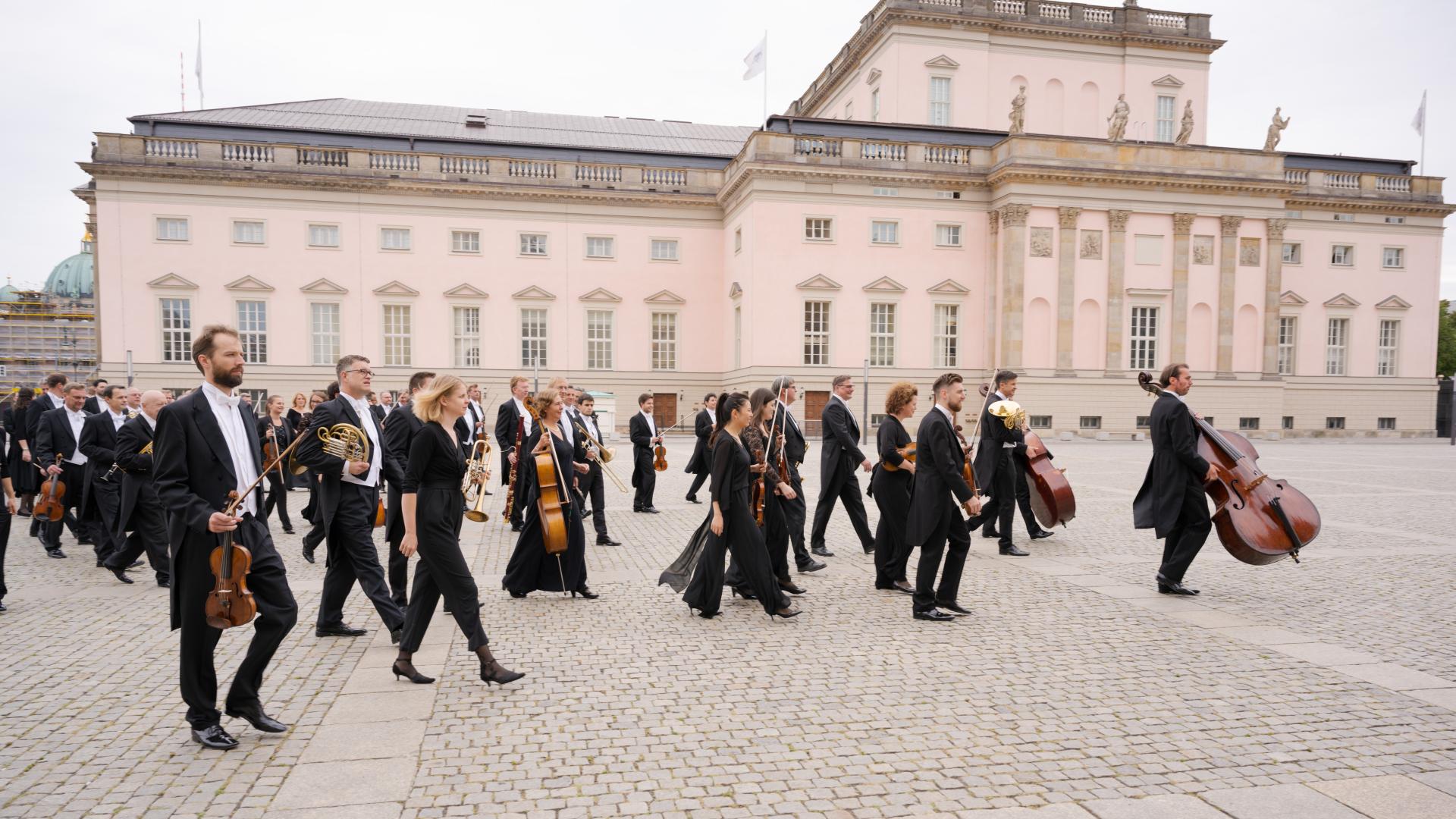 Das Orchester schreitet samt Instrumenten über einen Platz, im Hintergrund die Staatsoper Berlin.