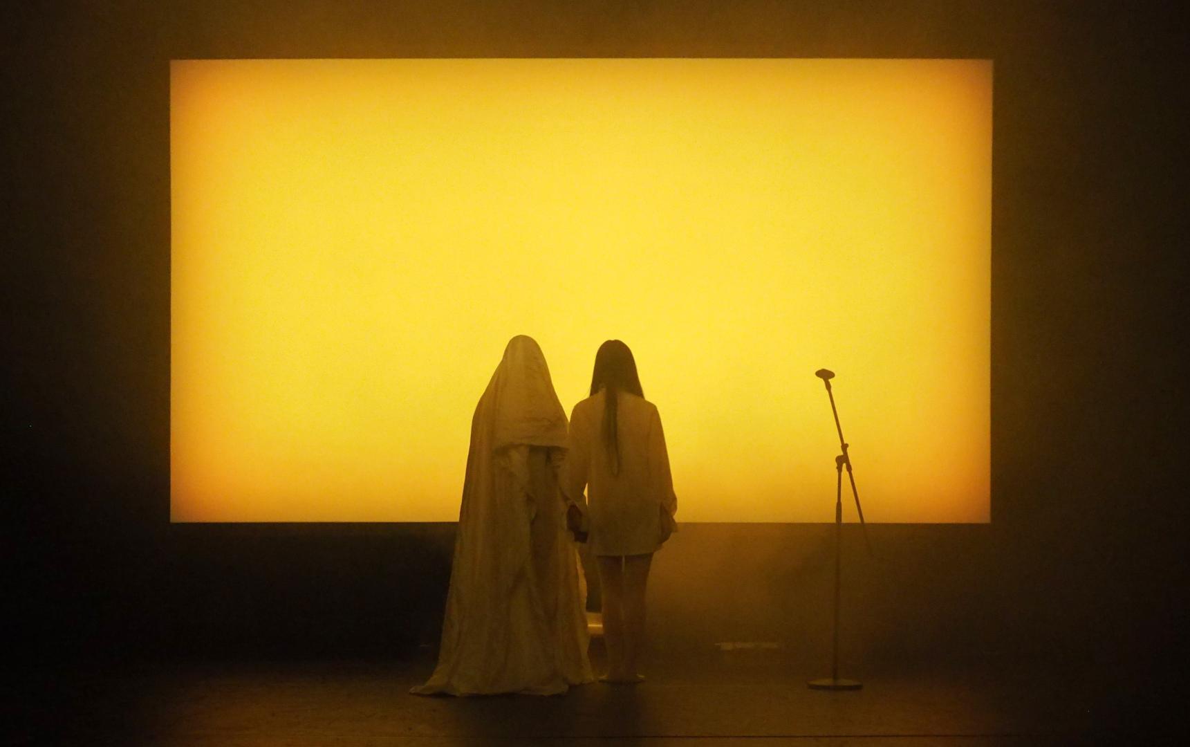 Zwei dem Publikum die Rücken zuwendenden Personen auf einer Bühne vor einer gelb leuchtenden Leinwand