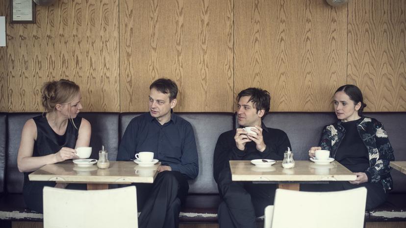 Die vier Mitglieder des Ensemble Adapter sitzen einer holzvertäfelten Bar und trinken Kaffee.