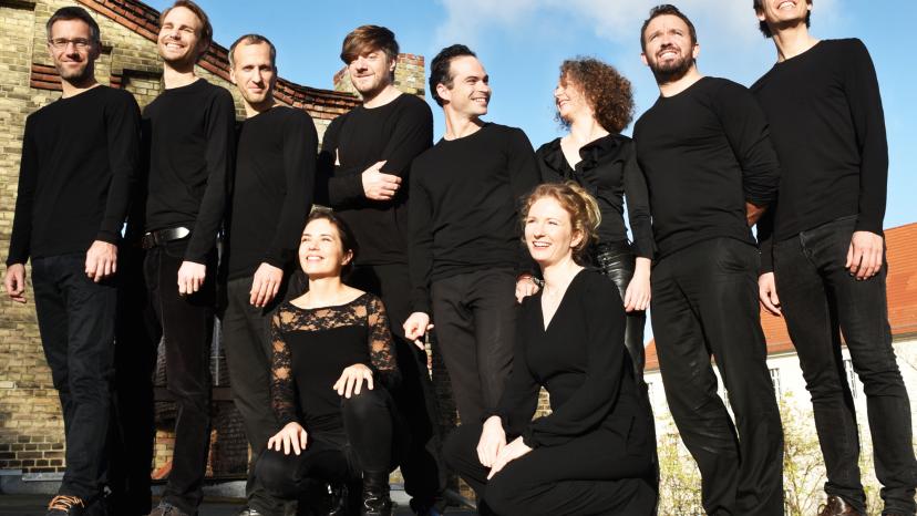 Die Mitglieder des Ensembles stehen auf dem Dach eines urbanen Backsteinhauses. Sie sind schwarz gekleidet und lächeln. 